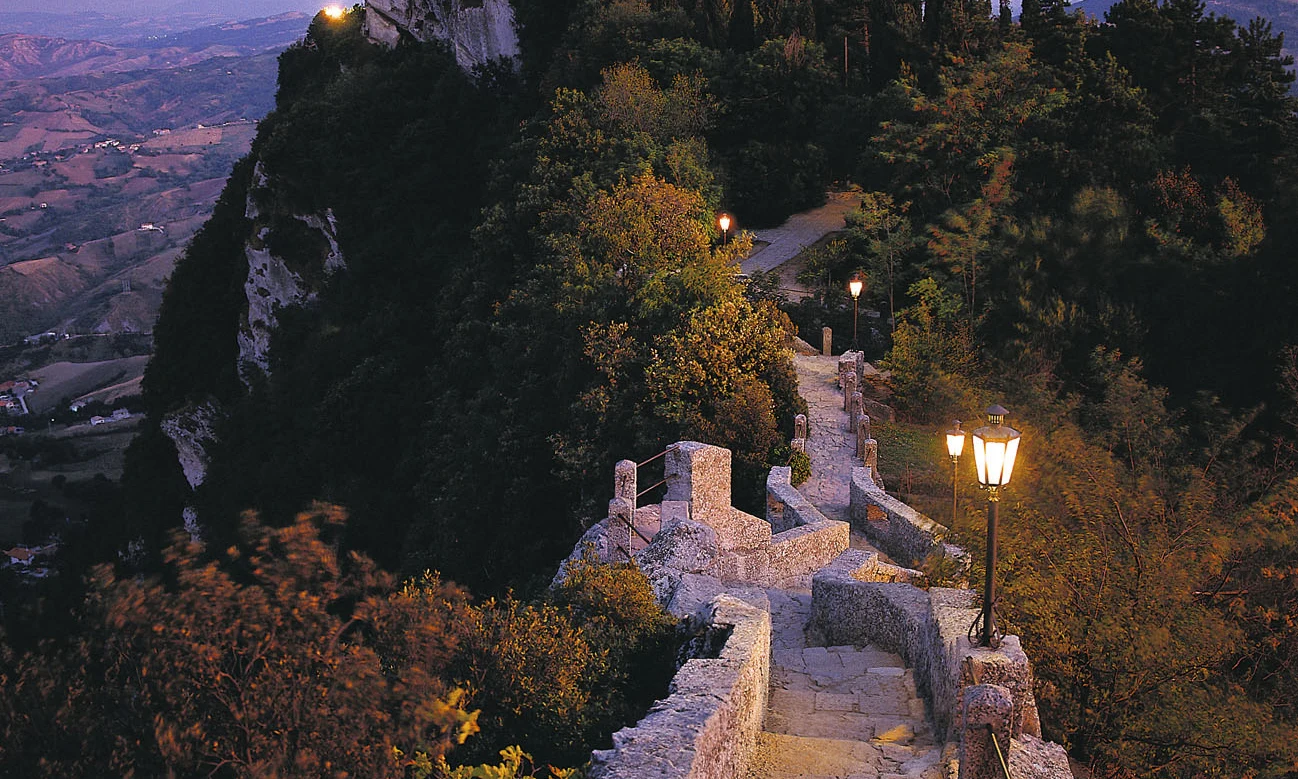 Castle sits atop a wooded, rocky hill, with a lamp-lit stone path winding upwards toward it, overlooking a valley at Centro Vacanze San Marino