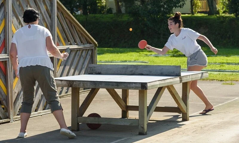 Two people playing table tennis outdoors, with a wooden structure and greenery in the background at Domaine de Mesqueau