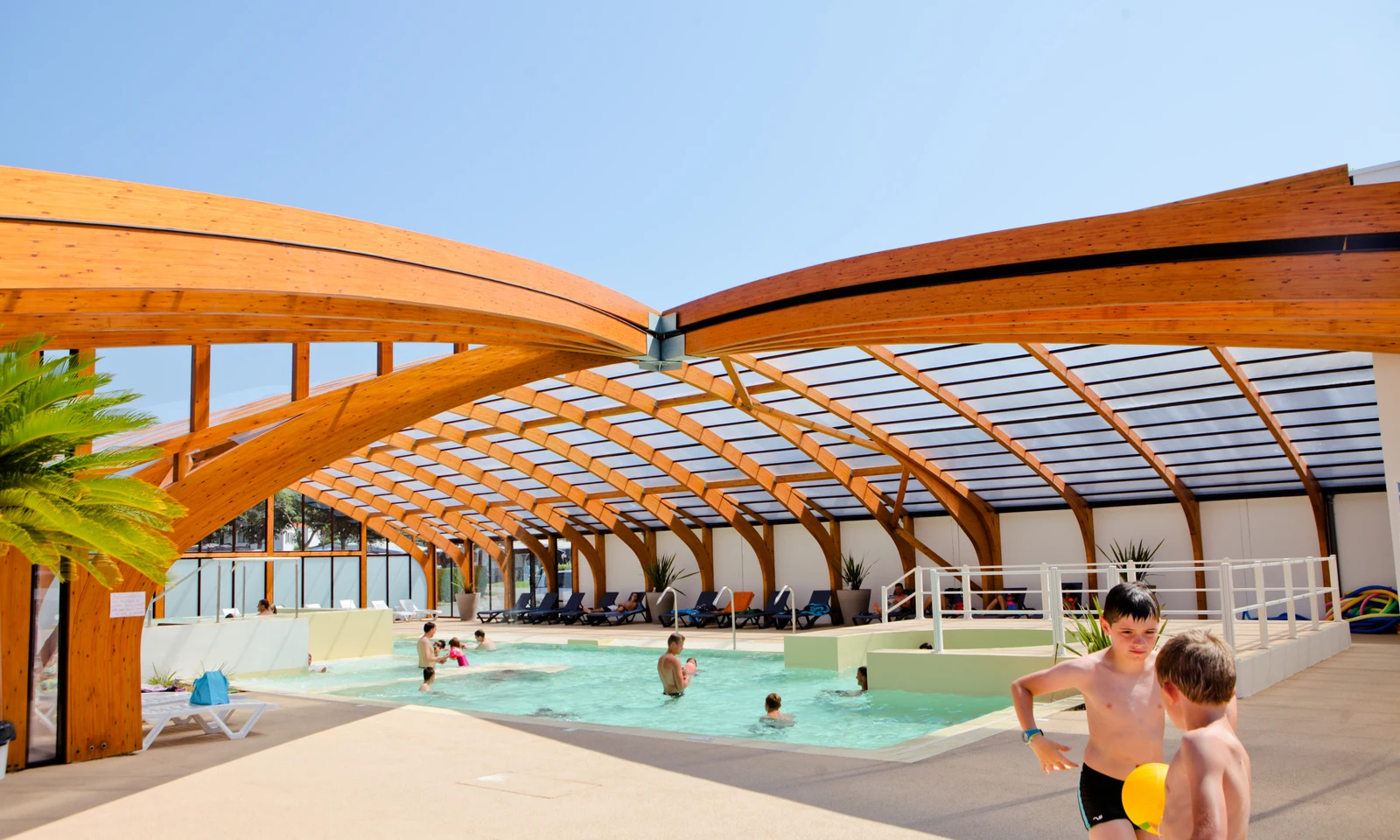 Children play in an indoor pool under a wooden arched roof, with lounge chairs and tropical plants around; at Ur-Onéa