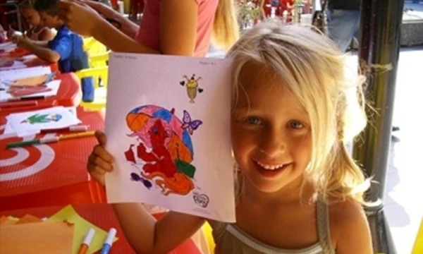 Child proudly holding colorful drawing, smiling at a table with art supplies, surrounded by people also drawing at Oasi