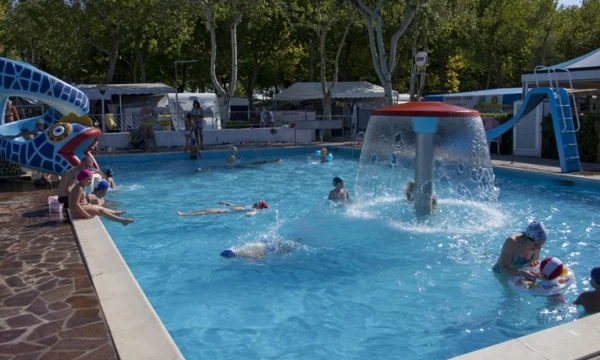 Children swim and play in a large outdoor pool with water features, surrounded by trees and camping amenities at Oasi