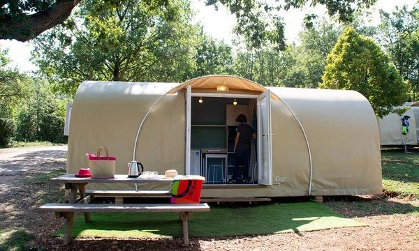 A person arranges items inside a beige tent-like structure beside a picnic table in a wooded area at de l'Aigle