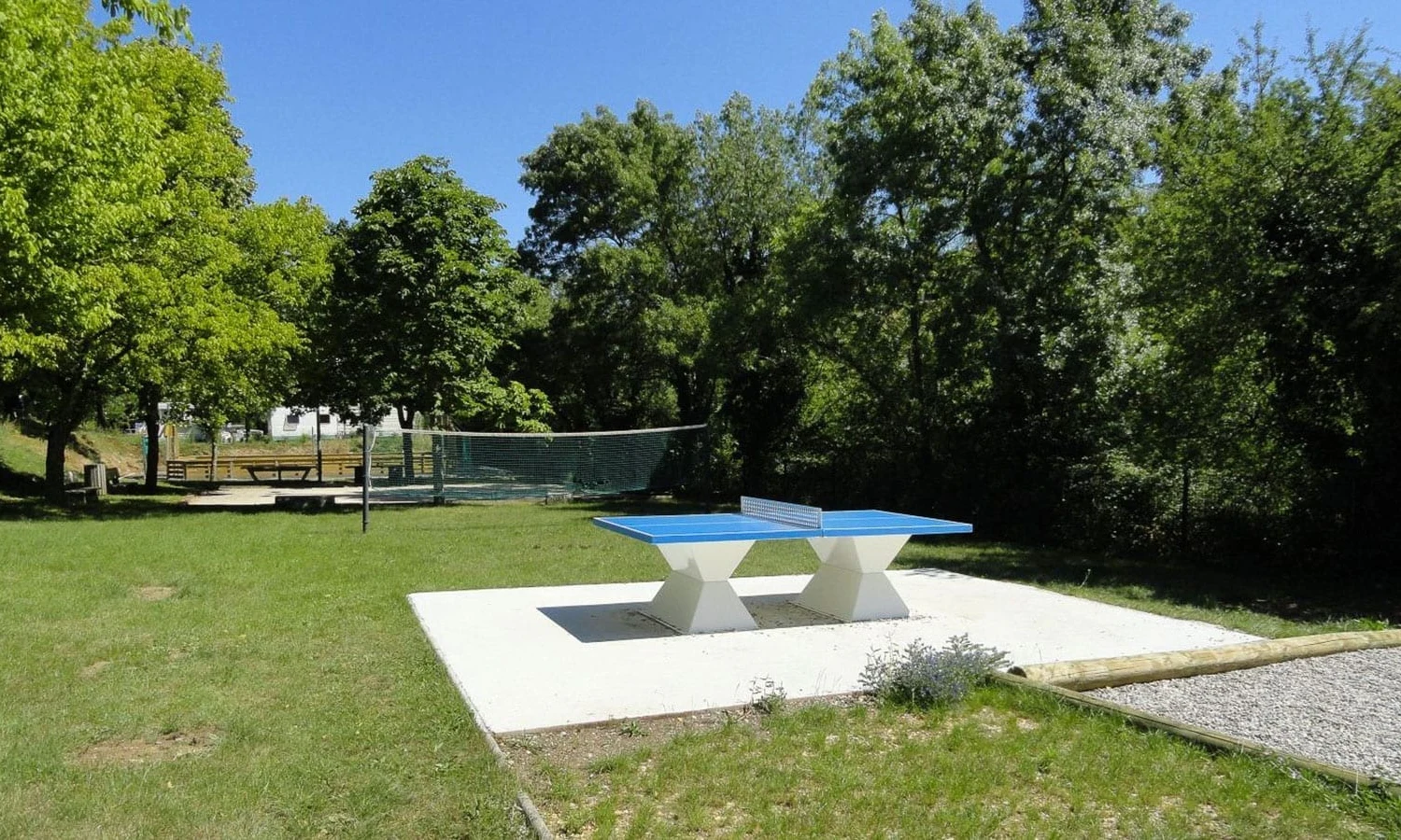 Ping pong table with a blue surface stationed on a concrete slab, surrounded by grassy field and trees at Ardeche Camping 