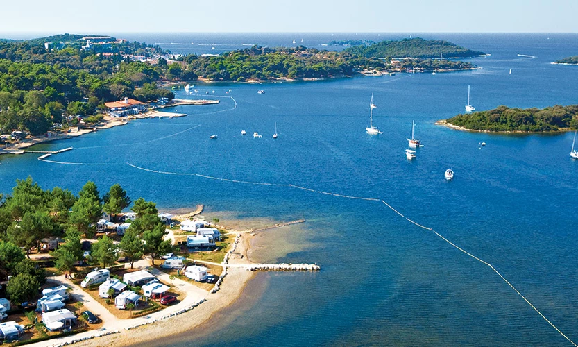 Campsite with RVs and tents by a forested coastline, adjacent to a calm sea with anchored sailboats at Orsera