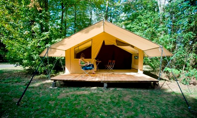 Large tent with elevated deck, person sitting on chair reading, surrounded by a lush forest at de l'Aigle