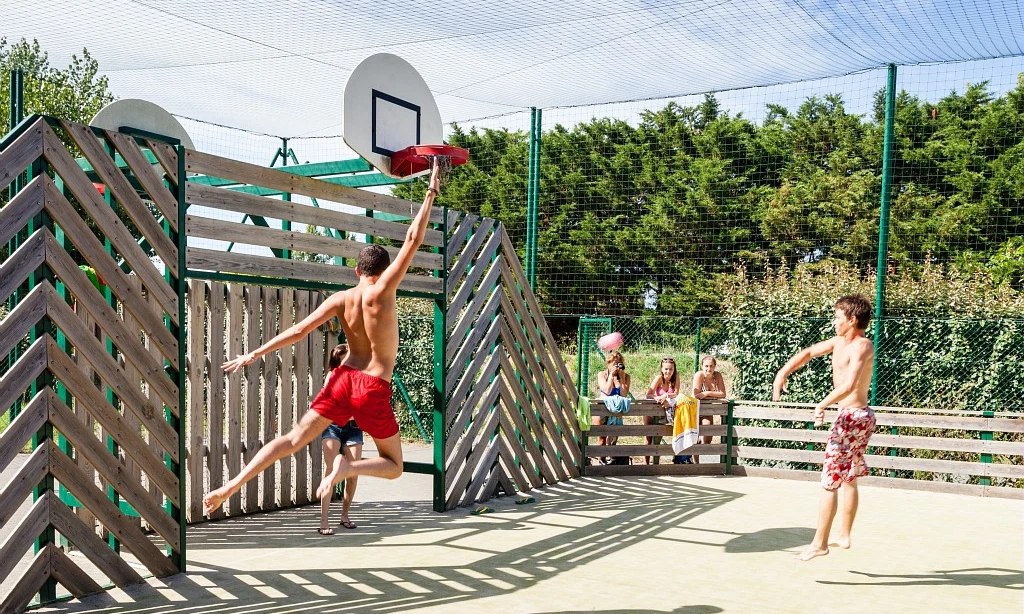 Two boys playing basketball, with one jumping to shoot; spectators watch from a bench in an enclosed court at Les Amiaux