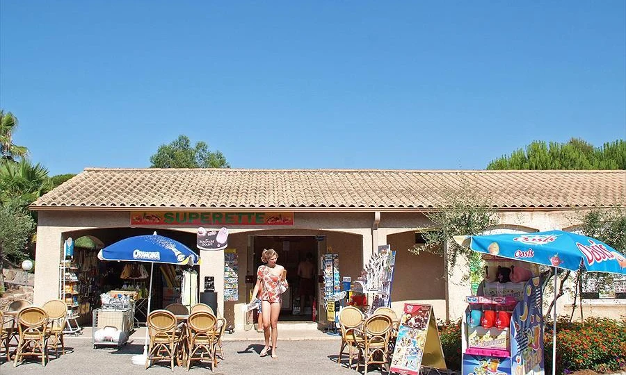 A person exits a small market under a clear sky, surrounded by colorful umbrellas and outdoor seating at Leï Suves