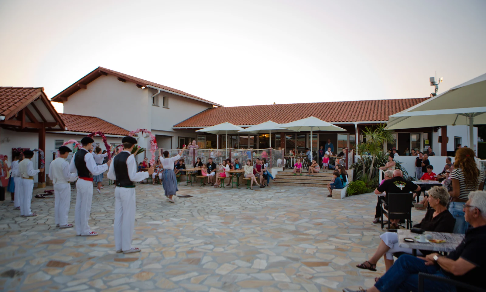 Dancers in traditional attire perform outdoors for seated audience near a building with red-tiled roofs at Ur-Onéa