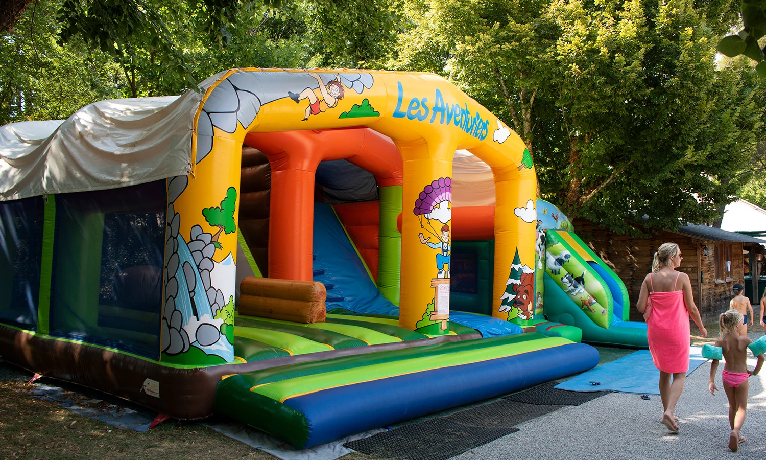 Inflatable play structure with slides and climbing features surrounded by trees, and people walking nearby at Ardeche Camping 