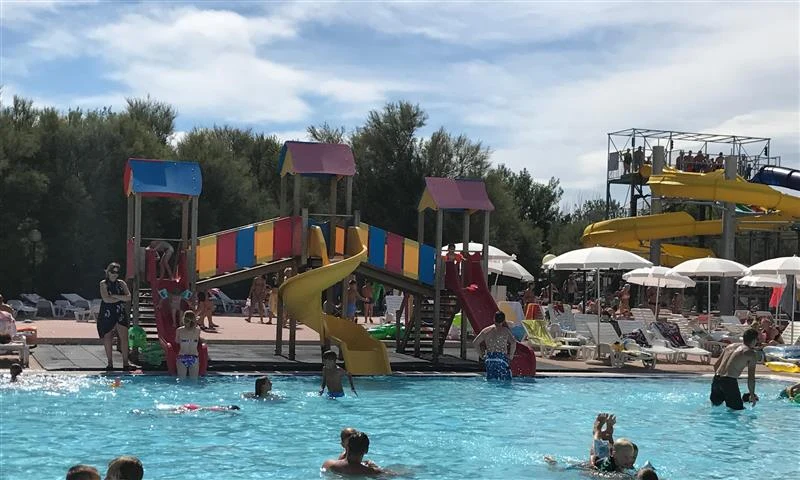 Children play on colorful slides and swim in a crowded pool, surrounded by trees and lounge chairs at Isamar