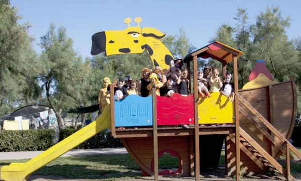 Colorful wooden playground structure resembling a giraffe, with children playing on it, in a grassy, tree-lined park at Oasi