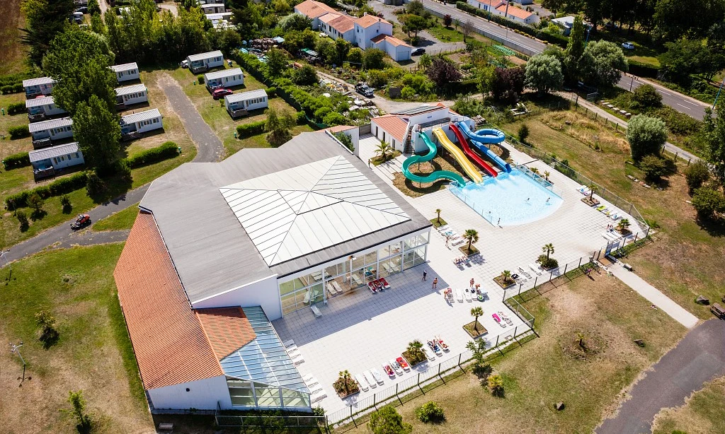 Aerial view of a recreational building with a water park and slides surrounded by holiday cabins and trees at Les Amiaux