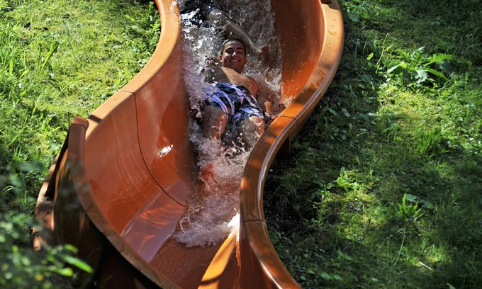 Person sliding down a curved, wet waterslide surrounded by green grass and foliage, enjoying summertime activities at Natterer See