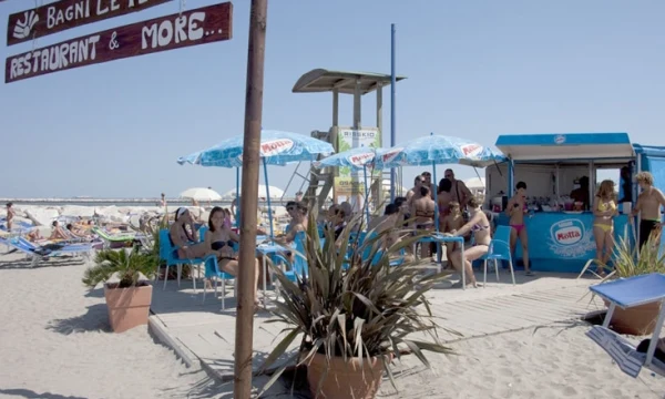 People sitting under umbrellas at outdoor beach café, with sand, plants, and a signpost in the background at Oasi