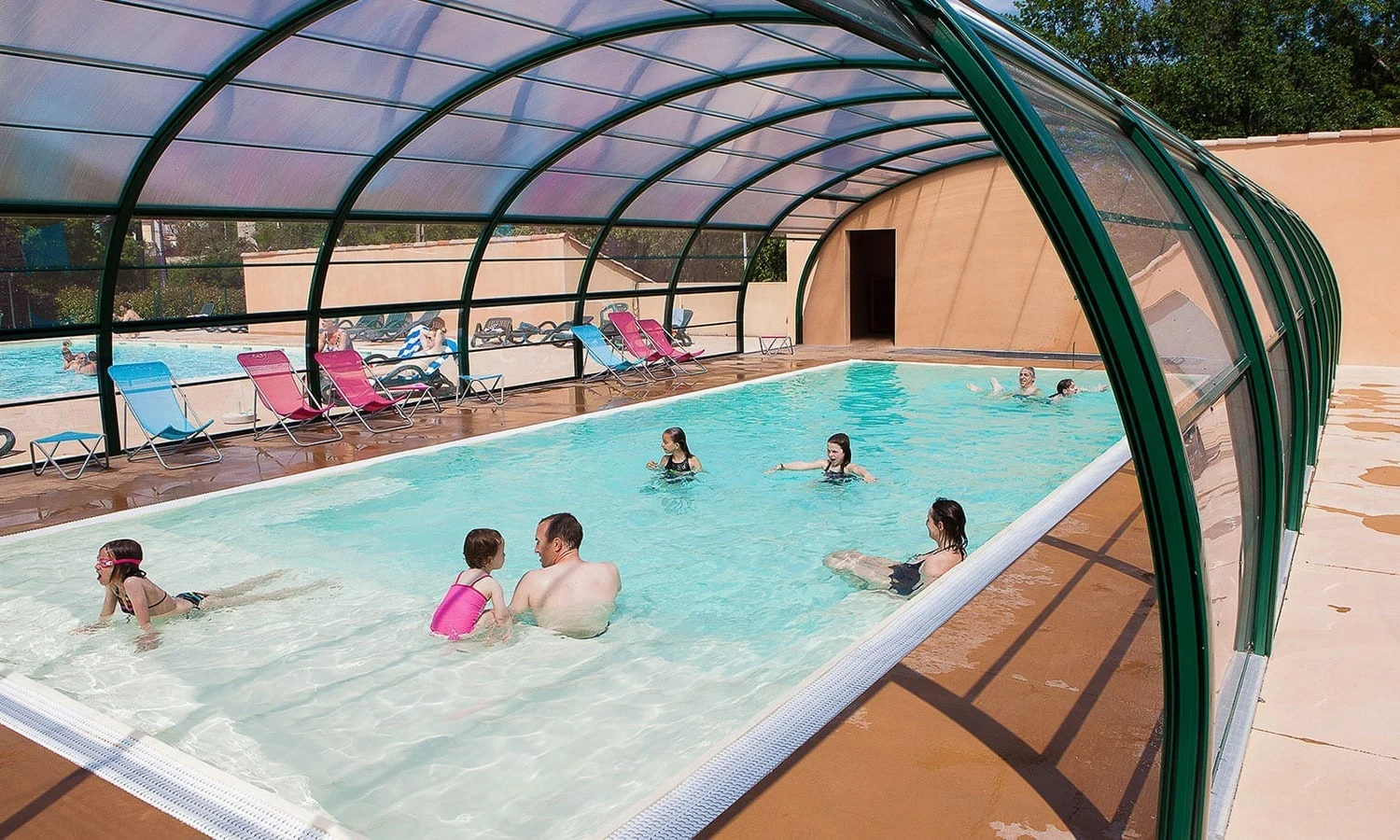 People swimming and lounging beside an enclosed pool with colorful deck chairs on a sunny day at Ardeche Camping 