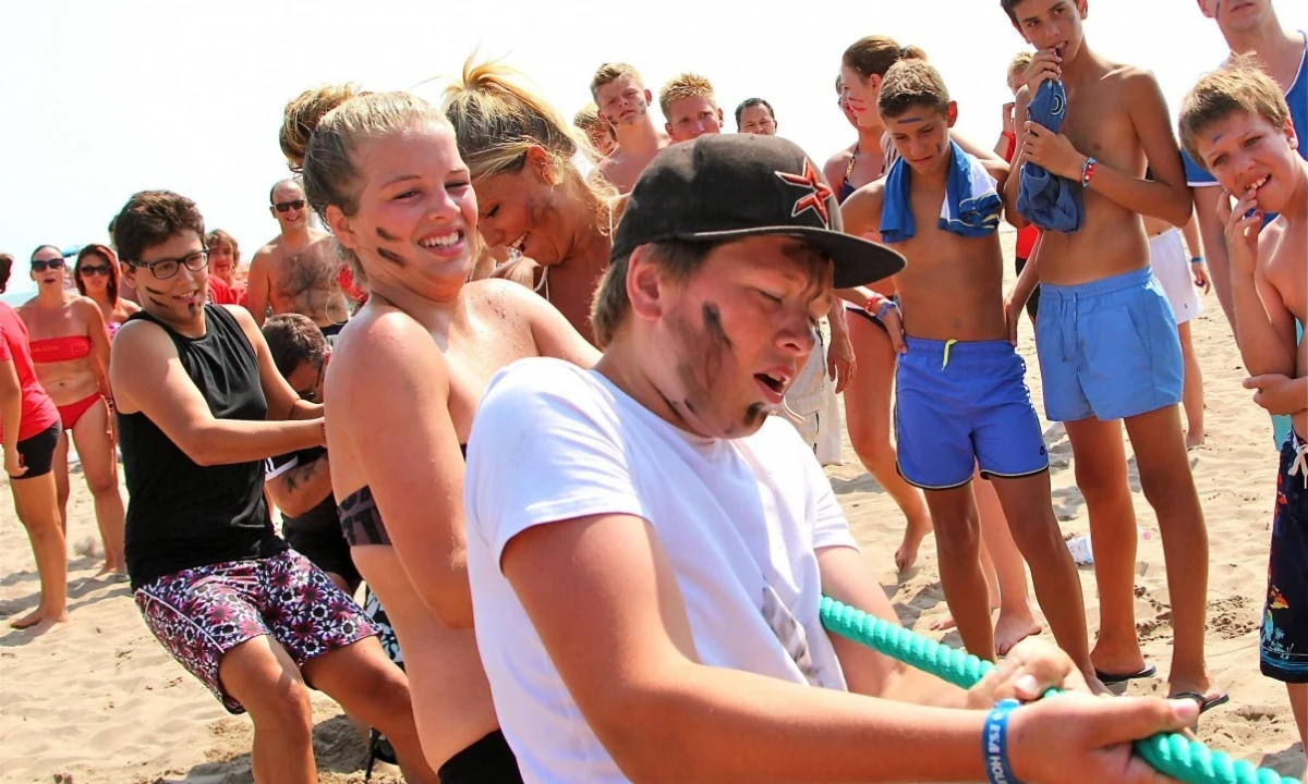 A group of teenagers pulls a rope in a tug-of-war on a sandy beach, with other campers watching at Isamar