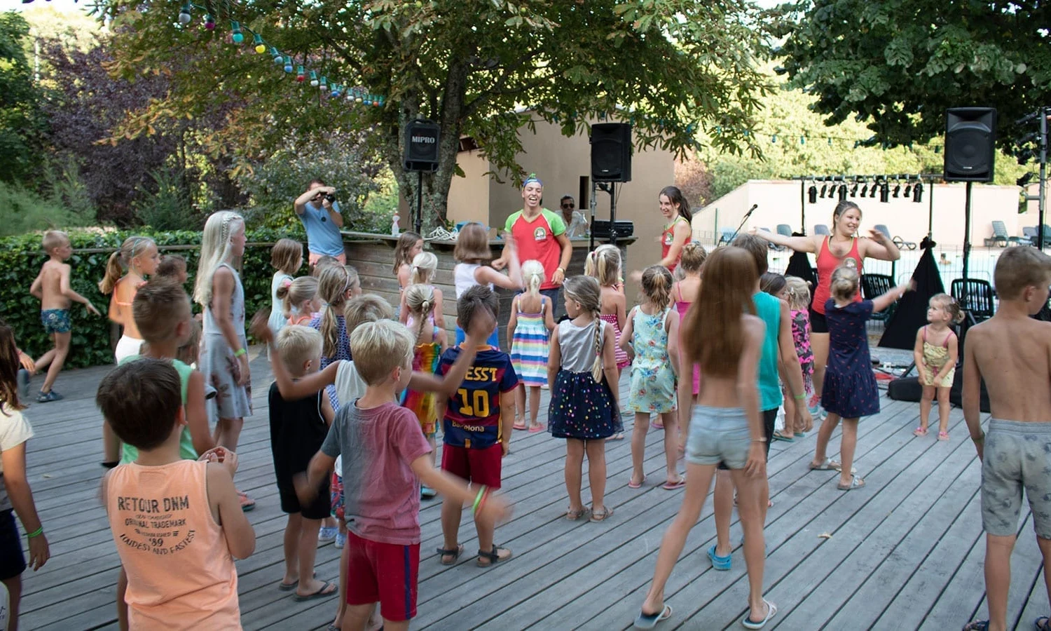 Children are dancing with instructors on a wooden deck under trees, with colorful fairy lights overhead at Ardeche Camping 