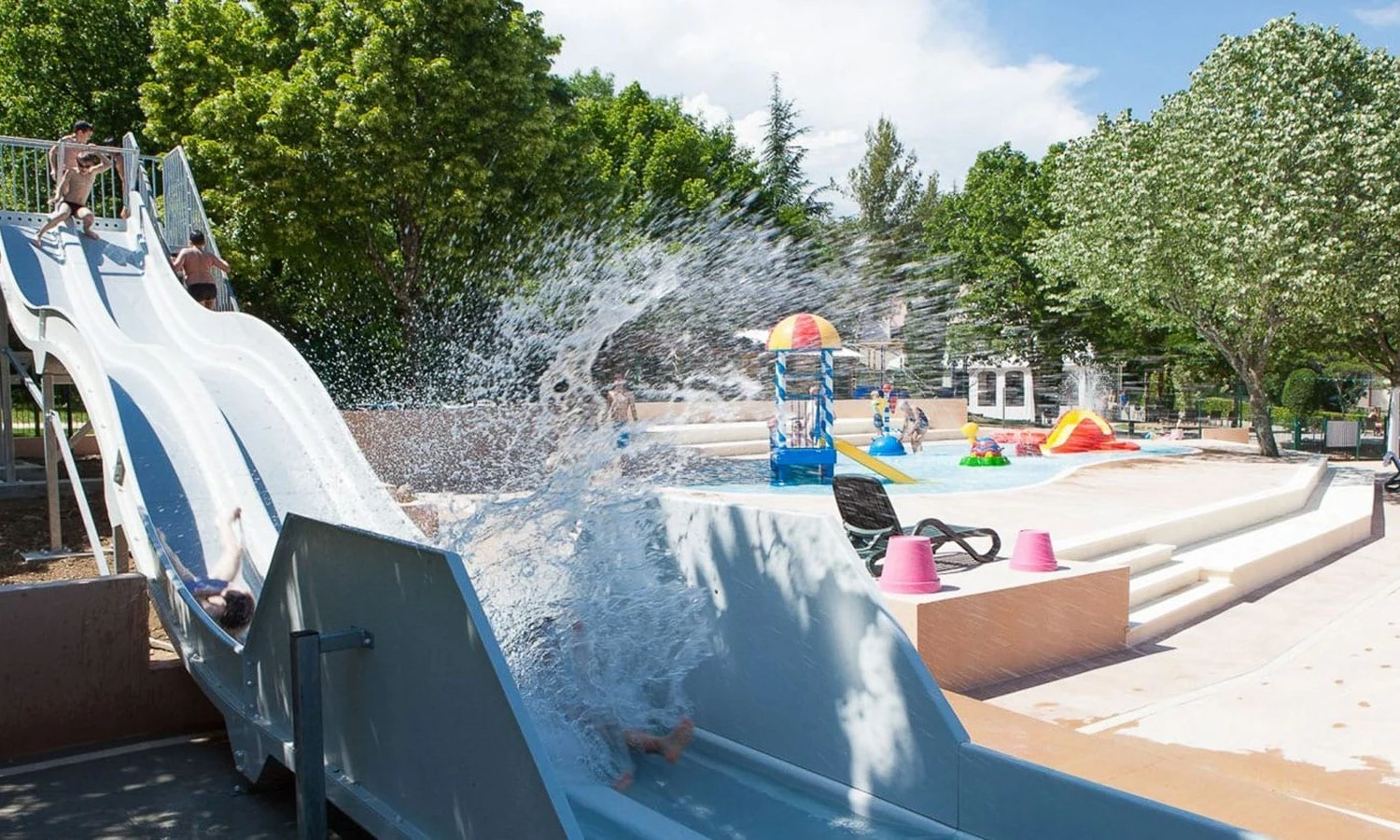 Waterslide splashes children into shallow pool, framed by trees and colorful play structures, under a bright sky at Ardeche Camping 