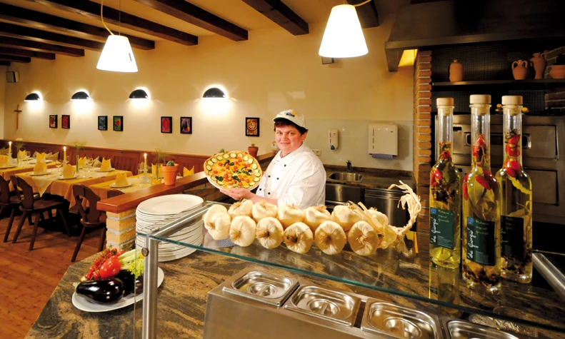 Chef presenting a freshly made pizza in a decorated restaurant kitchen, with tables set for dining; at Natterer See