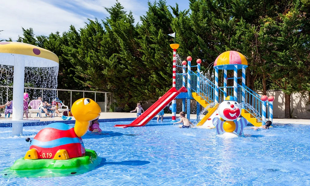 Kids playing on colorful water slides and animal-themed structures in a pool area surrounded by trees at Les Amiaux