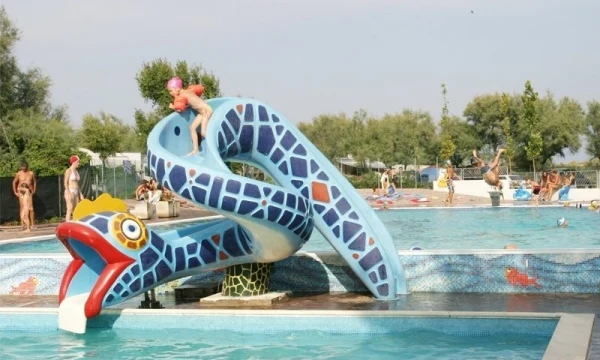 Child slides down a colorful snake slide into the swimming pool, surrounded by other children and adults at Oasi