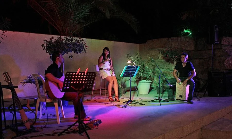 Musicians perform with guitar and percussion under vibrant lights on an outdoor stage surrounded by plants at Leï Suves