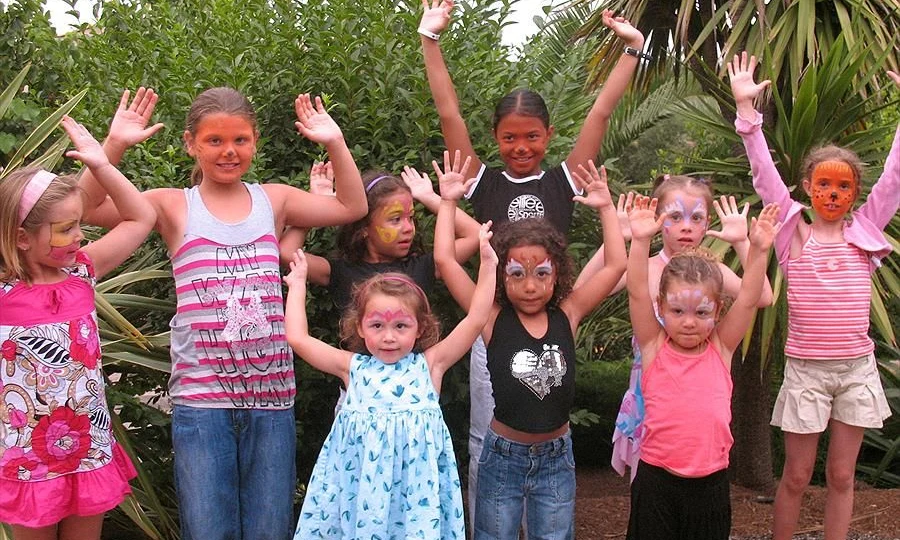 Children with painted faces stand under raised hands, smiling together, amidst lush greenery at Leï Suves