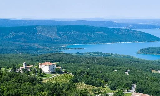 A grand villa overlooks lush forests and a tranquil lake, framed by distant rolling mountains at de l'Aigle