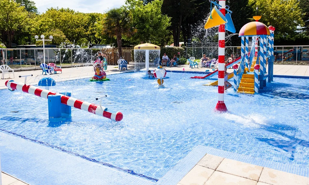 Children's water playground with colorful structures, spouting fountains, and slides, surrounded by trees and lounge chairs at Les Amiaux