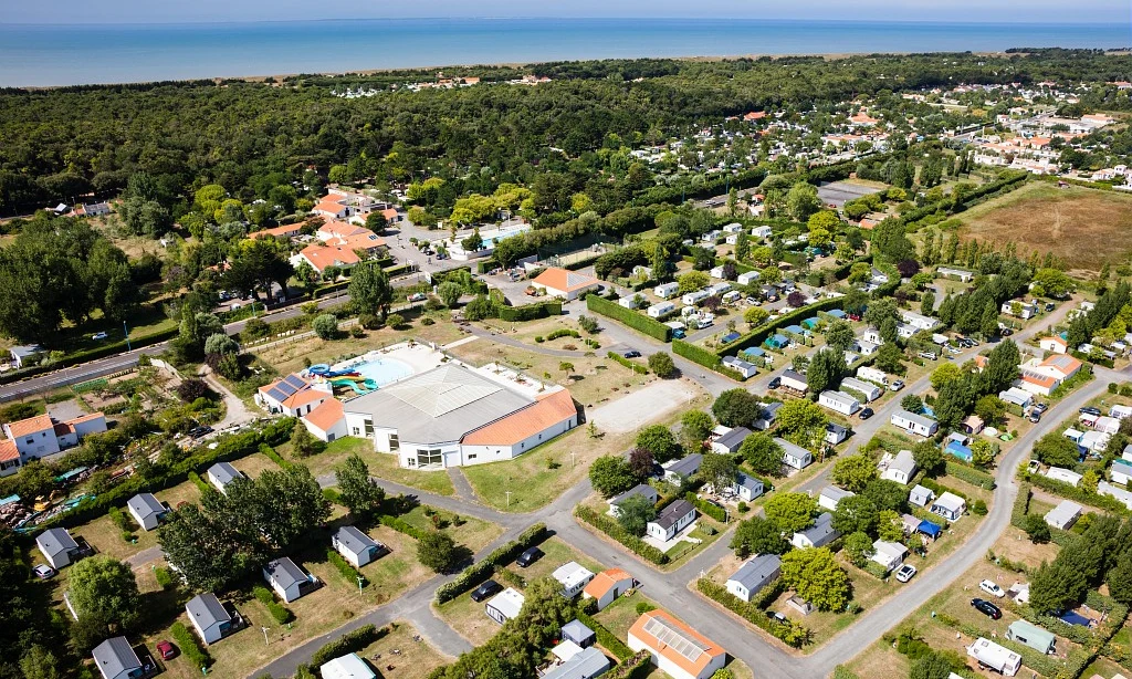 Aerial view of a campground with cabins and RVs, featuring a central building with a pool near a forest and beach at Les Amiaux