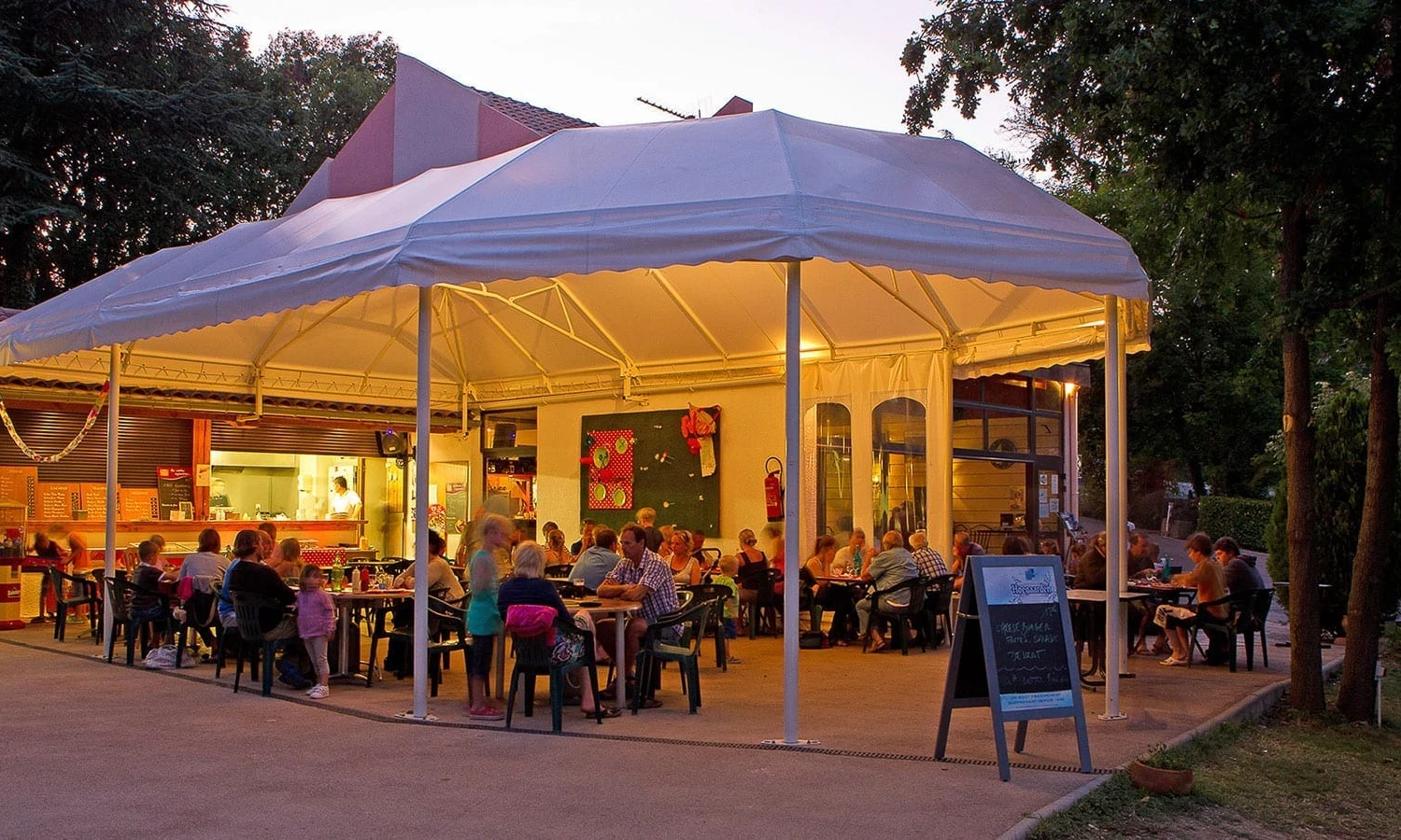 People dining under a lit canopy at an outdoor restaurant, surrounded by trees and a soft evening light at Ardeche Camping 