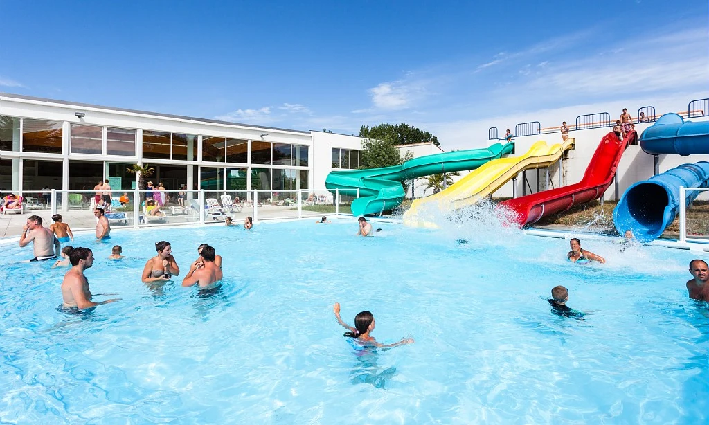 People swimming in a pool with water slides, surrounded by a building with large windows and lounging area at Les Amiaux