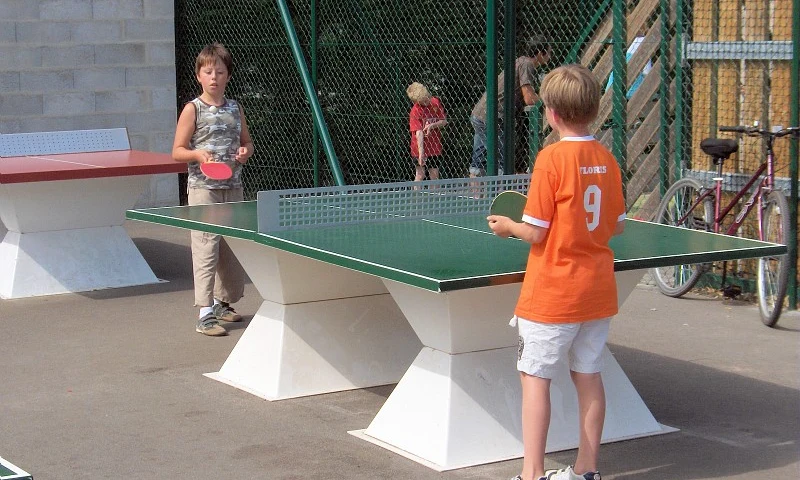 Two children playing table tennis outside near a fence and bench at Les Amiaux