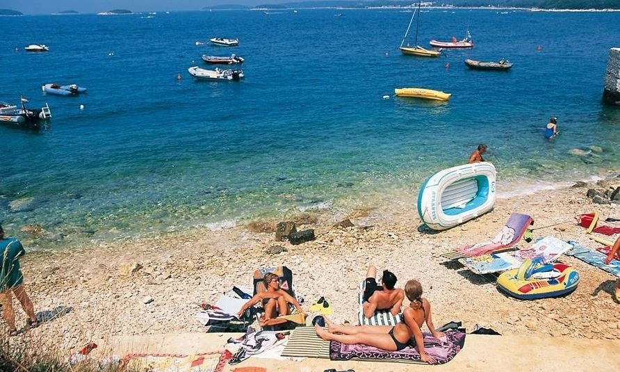 People lounging on a pebble beach with inflatable boats, near a calm, blue sea at Orsera