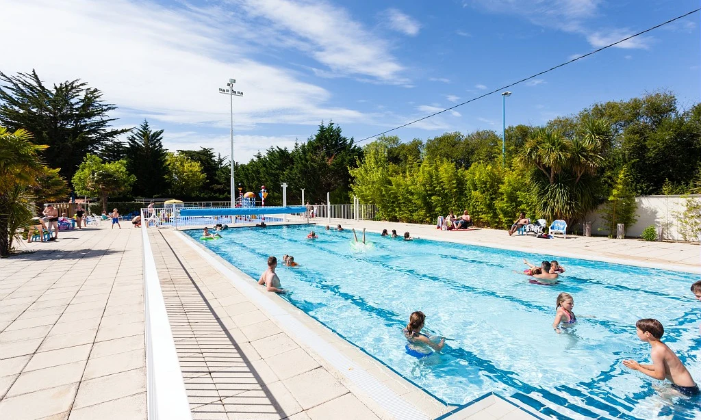 People swimming and lounging by a long outdoor pool surrounded by trees and lounge chairs at Les Amiaux