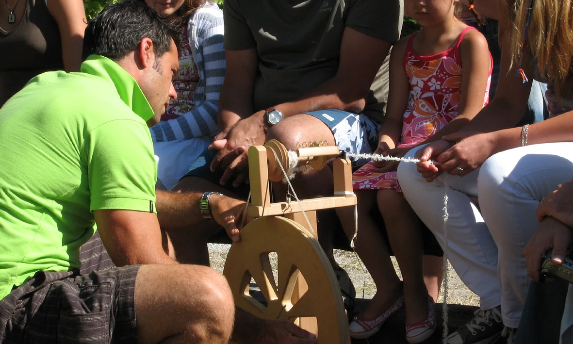 A person spins wool into yarn using a spinning wheel, surrounded by onlookers in an outdoor setting at Ur-Onéa