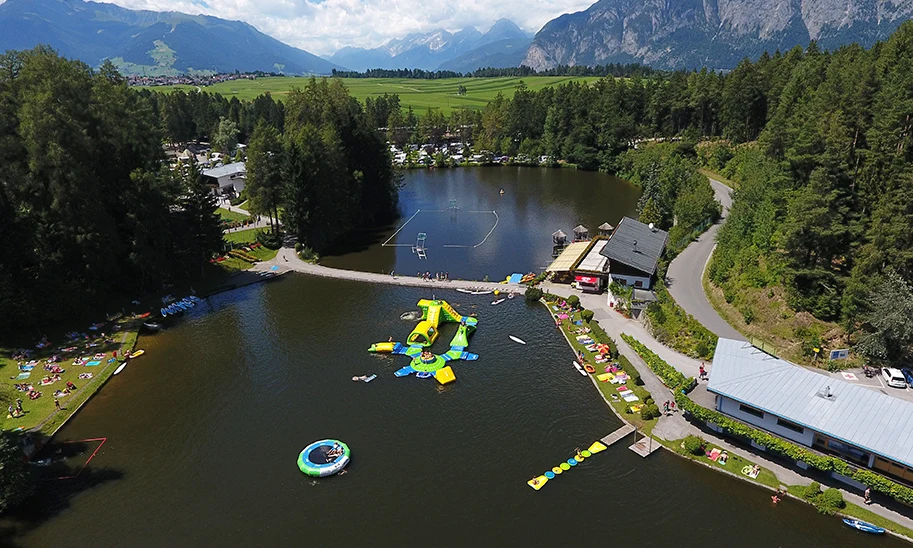 An inflatable water park floats on a lake surrounded by trees and mountains on a sunny day at Natterer See