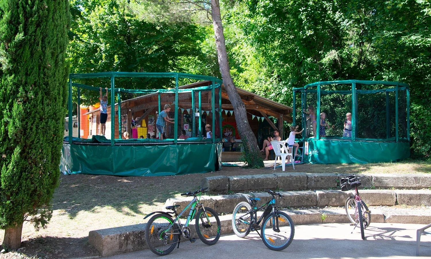 Children jumping on trampolines near a wooden structure; adults relax in chairs beside bicycles and trees at Ardeche Camping 