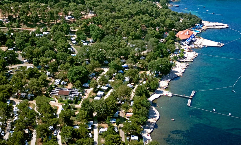 Aerial view of a campsite with numerous caravans surrounded by dense trees near a shoreline with clear blue water at Orsera