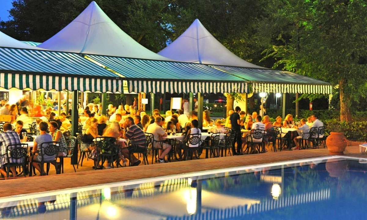 People dining under a large, striped canopy by a swimming pool, surrounded by trees at Isamar