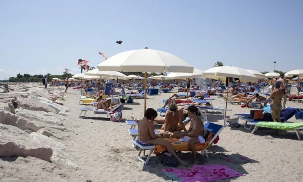 Sunbathers relaxing on lounge chairs under beach umbrellas on a sandy shoreline with some people building a sandcastle, at Oasi