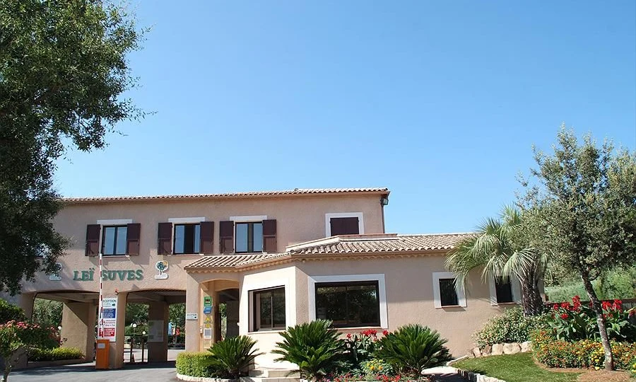 A beige building with brown shutters stands amidst lush greenery and flowers at Leï Suves