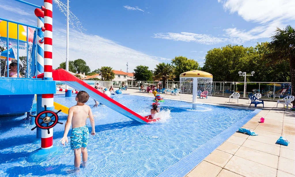 Children playing on a waterslide in a bright, blue pool with a splash feature under sunny skies at Les Amiaux