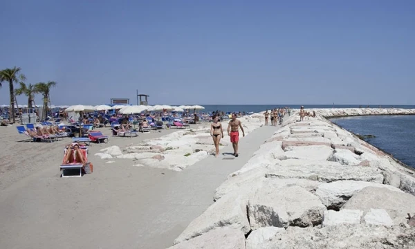 People walking on a stone pier, with a beach lined with sunbathers and umbrellas, meeting the ocean horizon at Oasi