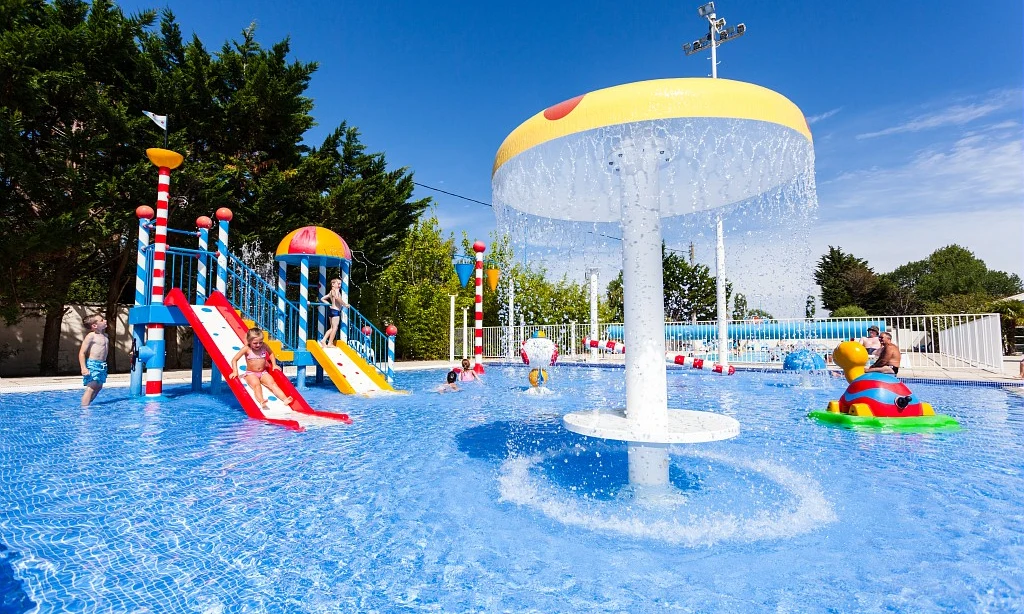 Children playing on colorful water slides and under a large water sprinkler in a pool area, surrounded by trees at Les Amiaux