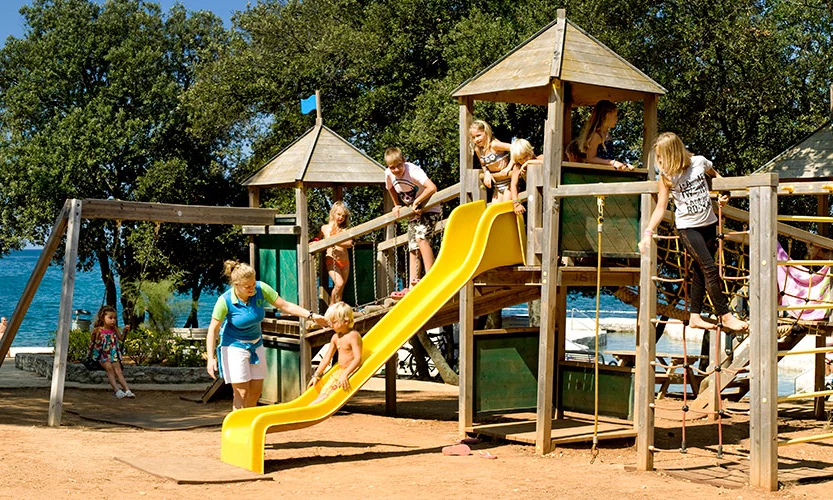 Children playing, climbing, and sliding on a wooden playground set by the beach, surrounded by trees at Orsera