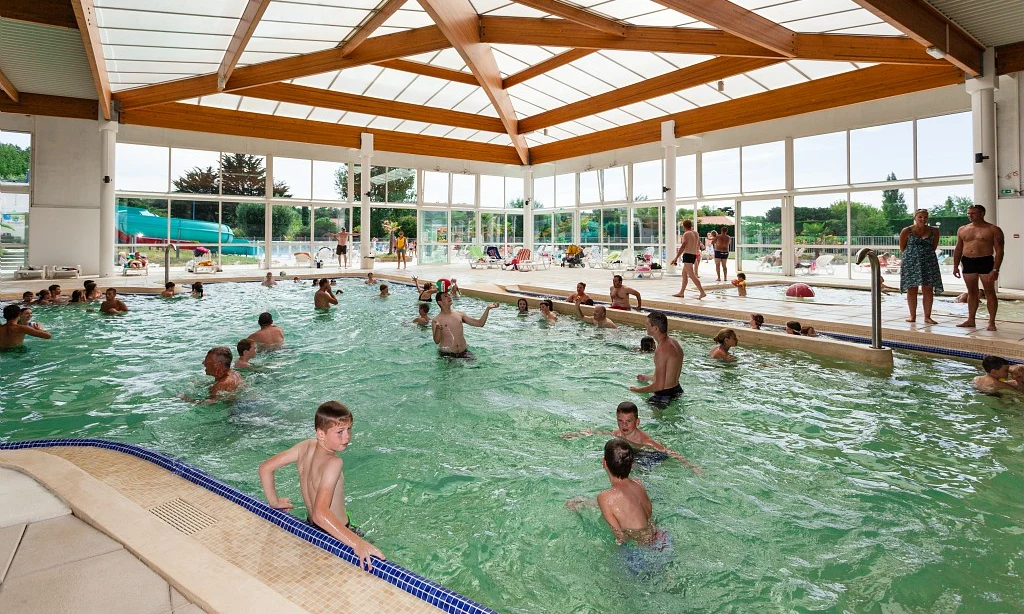 People swimming in an indoor pool, with large windows and a wooden ceiling at Les Amiaux