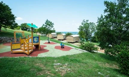 Playground structures sit on a colorful rubber mat in a grassy area with trees and wooden cabins nearby at de l'Aigle