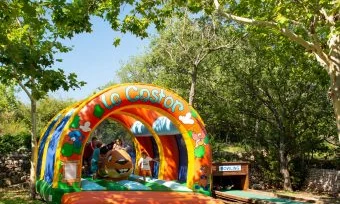 Children bounce inside a colorful inflatable castle under shaded trees; picnic tables and forested area in the background at de l'Aigle