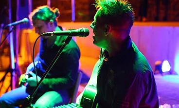 Two musicians playing guitars and singing into microphones on a dimly lit stage at Orsera